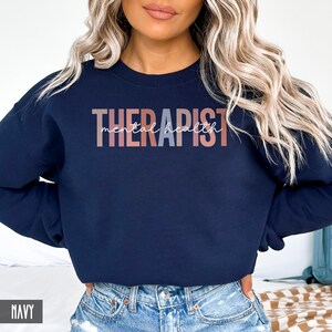 May include: A navy blue sweatshirt with the text "THERAPIST mental health" in a gradient of pink, peach, and gray.