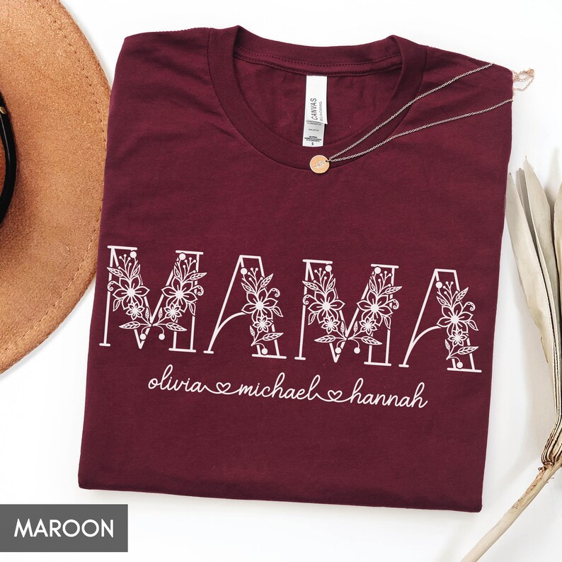 May include: Maroon t-shirt with the word "MAMA" in white floral lettering. Below the word "MAMA" are the names "olivia", "michael", and "hannah". A gold pendant necklace and a brown hat are visible.
