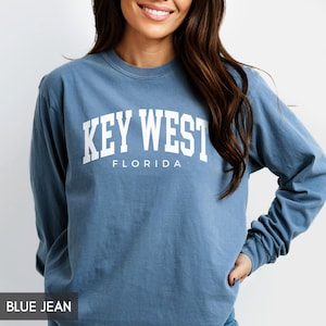 May include: Blue jean long-sleeve shirt with the text "KEY WEST FLORIDA" in white block letters. The shirt is a solid blue color and has a crew neck. The shirt is made of a soft, comfortable material.