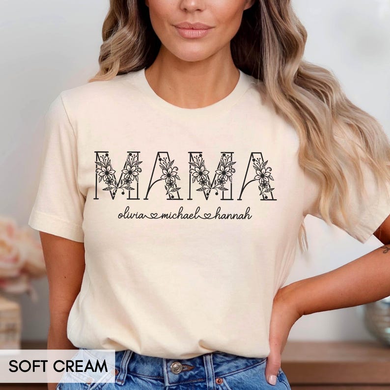 May include: Soft cream-colored t-shirt with the word "MAMA" in black, floral-filled letters. Below the word "MAMA" are the names Olivia, Michael, and Hannah, each with a heart symbol. The shirt is made of soft material.