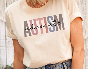 May include: A soft cream-colored short-sleeve t-shirt featuring the word AUTISM printed in large, blocky, multi-colored letters in shades of grey, pink, and blue, with the word advocate written in black cursive script across the center.