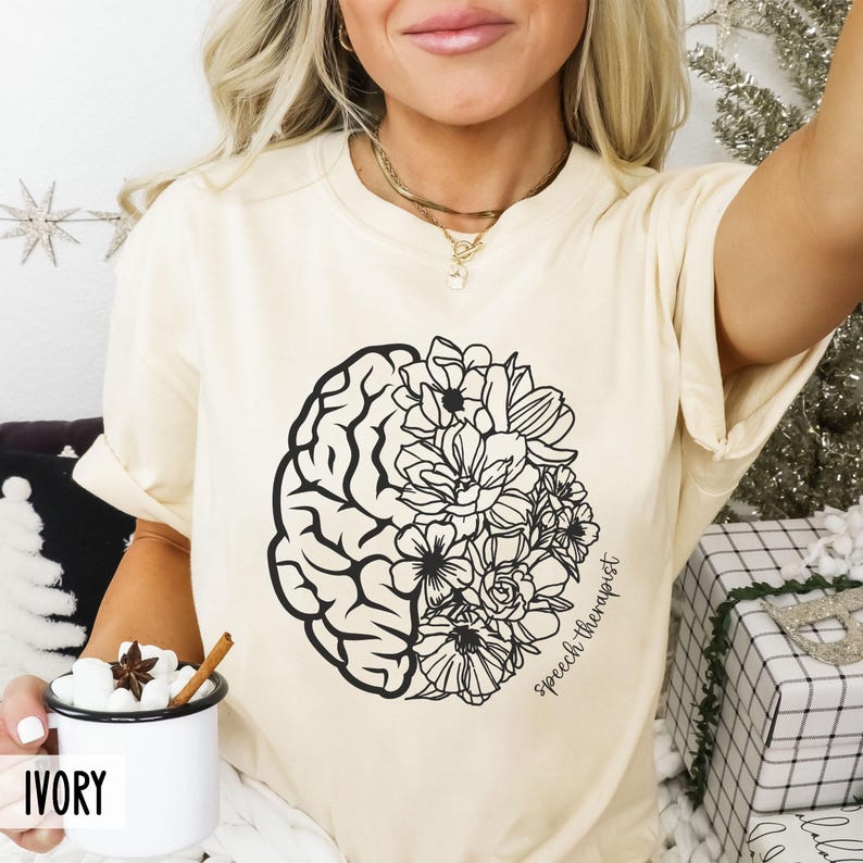 May include: A cream-colored t-shirt with a black and white design of a brain and flowers. The text "speech therapist" is written on the shirt.
