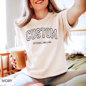 May include: A white cropped t-shirt with the text "CUSTOM" in a black varsity-style font. The text "OPTIONAL 2ND LINE" is printed below in a smaller font. The shirt is shown on a model wearing it with a coffee drink in hand. The color of the shirt is ivory.