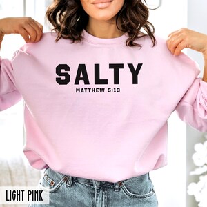 May include: A light pink sweatshirt with the text "SALTY" in black, followed by "MATTHEW 5:13" in smaller black text.