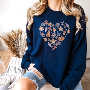 May include: A navy blue sweatshirt with a heart-shaped design featuring autumnal elements like leaves, acorns, pumpkins, and mushrooms. The design is in shades of orange, brown, and pink.