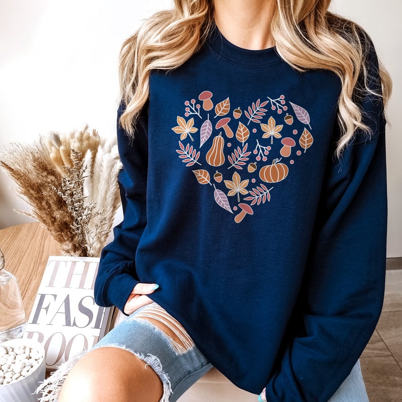 May include: A navy blue sweatshirt with a heart-shaped design featuring autumnal elements like leaves, acorns, pumpkins, and mushrooms. The design is in shades of orange, brown, and pink.