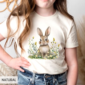 May include: A white t-shirt with a brown bunny rabbit surrounded by yellow and white flowers. The bunny is looking at the viewer. The shirt is labeled "NATURAL".