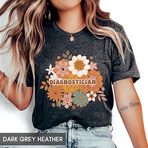 May include: Dark grey heather t-shirt with a floral design and the word "DIAGNOSTICIAN" in a retro font. The floral arrangement includes flowers in shades of orange, pink, white, and green. The shirt has a v-neck.