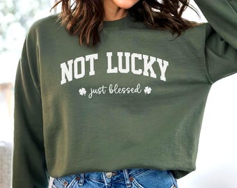 May include: A green sweatshirt with the text "NOT LUCKY just blessed" in white lettering. The sweatshirt is shown on a model wearing blue jeans.