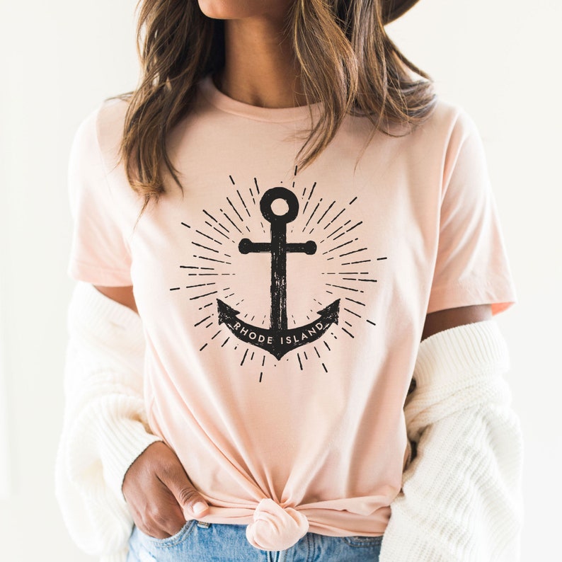 May include: A pink t-shirt featuring a black anchor graphic and the text "RHODE ISLAND". The anchor is surrounded by a sunburst design.