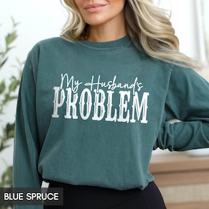 May include: A blue spruce long-sleeve crewneck sweatshirt featuring the white text My Husband's PROBLEM printed across the chest. The garment has a solid color finish with a ribbed neckline and cuffs, providing a simple and functional design.