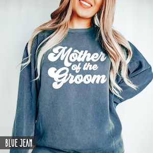 May include: Blue jean-colored sweatshirt with the text "Mother of the Groom" in white, retro-style lettering. The sweatshirt has long sleeves and a crew neck. The text "BLUE JEAN" is at the bottom of the image.