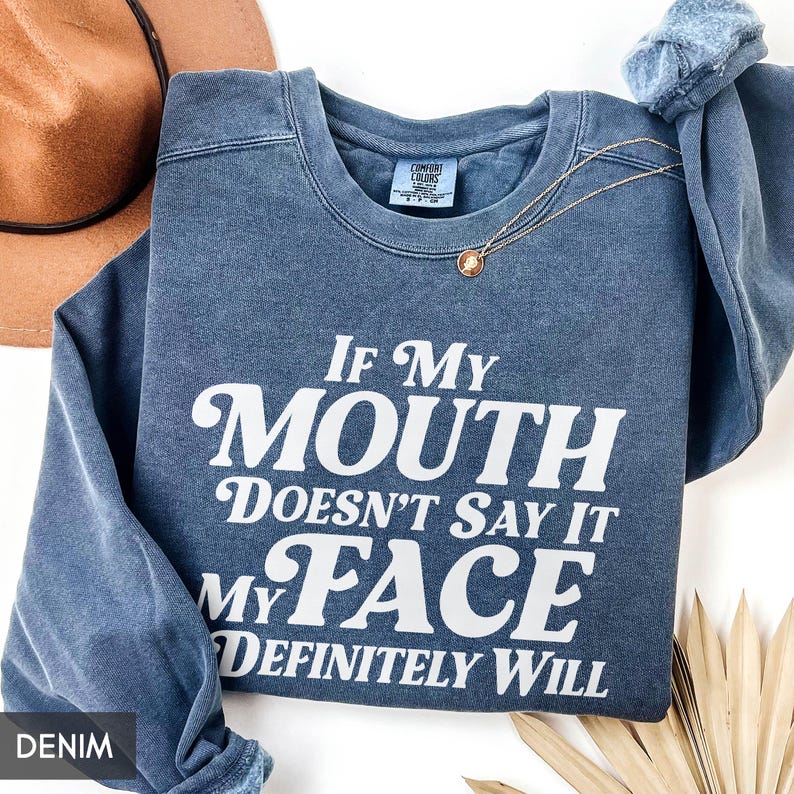 Puede incluir: Sudadera azul denim con el texto blanco "If My Mouth Doesn't Say It My Face Definitely Will". Un collar dorado con un colgante est&aacute; sobre la sudadera. Un sombrero marr&oacute;n es visible en el fondo.