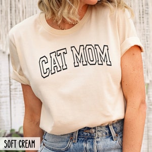 May include: A soft cream coloured t-shirt with the text "CAT MOM" in black outlined letters.