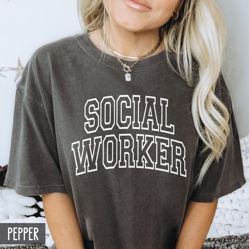 Puede incluir: Una camiseta gris oscuro con el texto "SOCIAL WORKER" impreso en blanco con un contorno blanco. El texto est&aacute; en una fuente de estilo universitario.