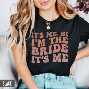 May include: A black t-shirt with the text "It's me, hi I'm the bride it's me" in a retro font with pink and orange color gradients. The text is centered on the shirt.