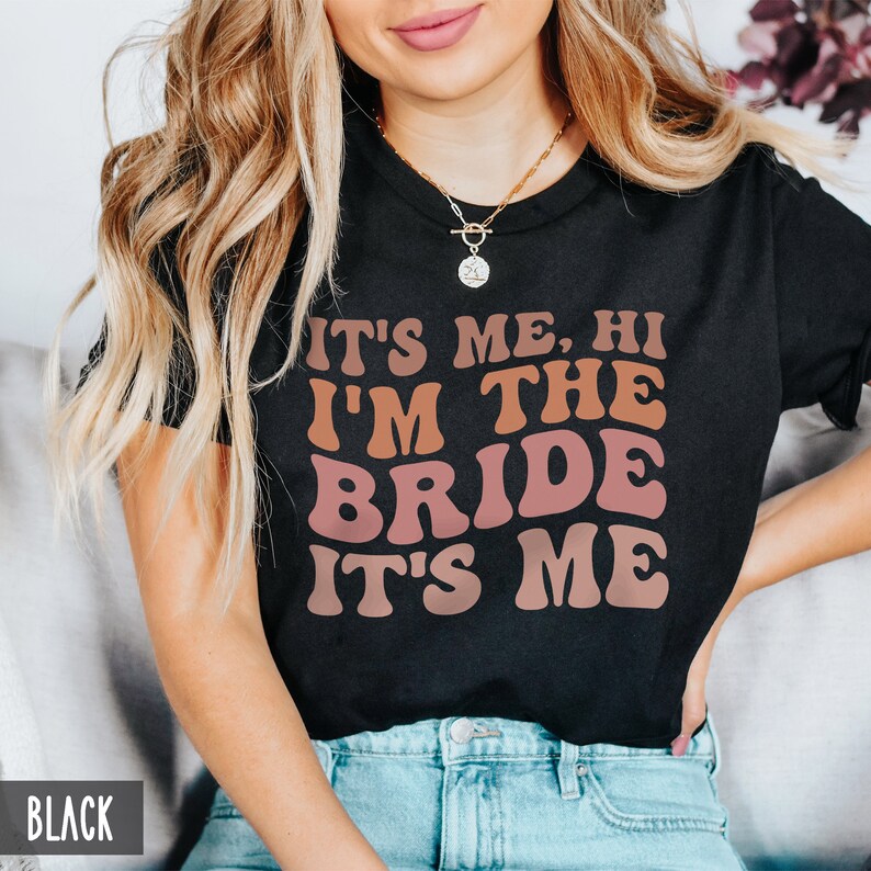 May include: A black t-shirt with the text "It's me, hi I'm the bride it's me" in a retro font with pink and orange color gradients. The text is centered on the shirt.