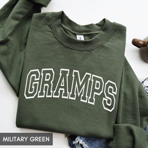 May include: A military green sweatshirt with the word "GRAMPS" in white, block letters. The sweatshirt is folded, with the sleeves tucked in. The text "MILITARY GREEN" is at the bottom of the image.