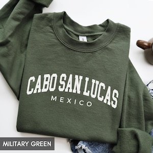 May include: Military green sweatshirt with the text "CABO SAN LUCAS MEXICO" in white block letters. The crew neck sweatshirt is folded and has a casual style.