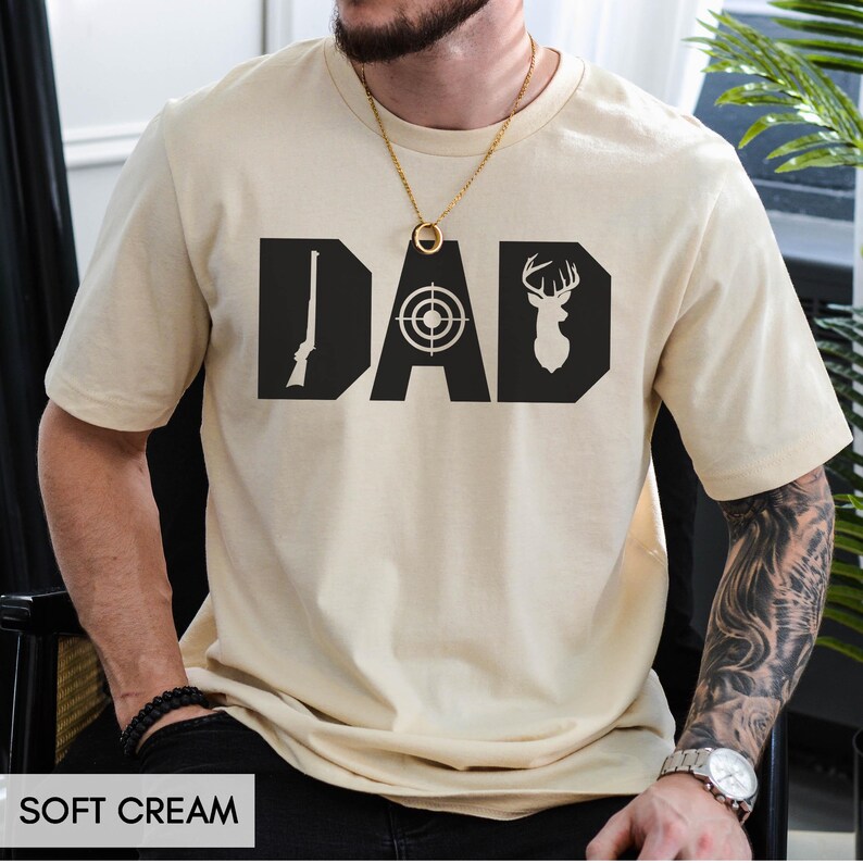 May include: Soft cream-colored t-shirt with the word "DAD" in bold black letters. The "D" features a rifle, the "A" a target, and the "D" a deer silhouette. The shirt is a casual wear item.
