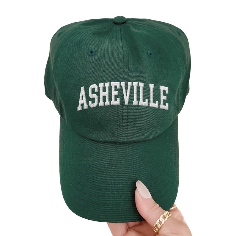 Puede incluir: Gorra de b&eacute;isbol verde oscuro con la palabra "ASHEVILLE" bordada en blanco en la parte delantera. La gorra tiene una visera curva y un bot&oacute;n en la parte superior. El sombrero est&aacute; hecho de una tela suave.