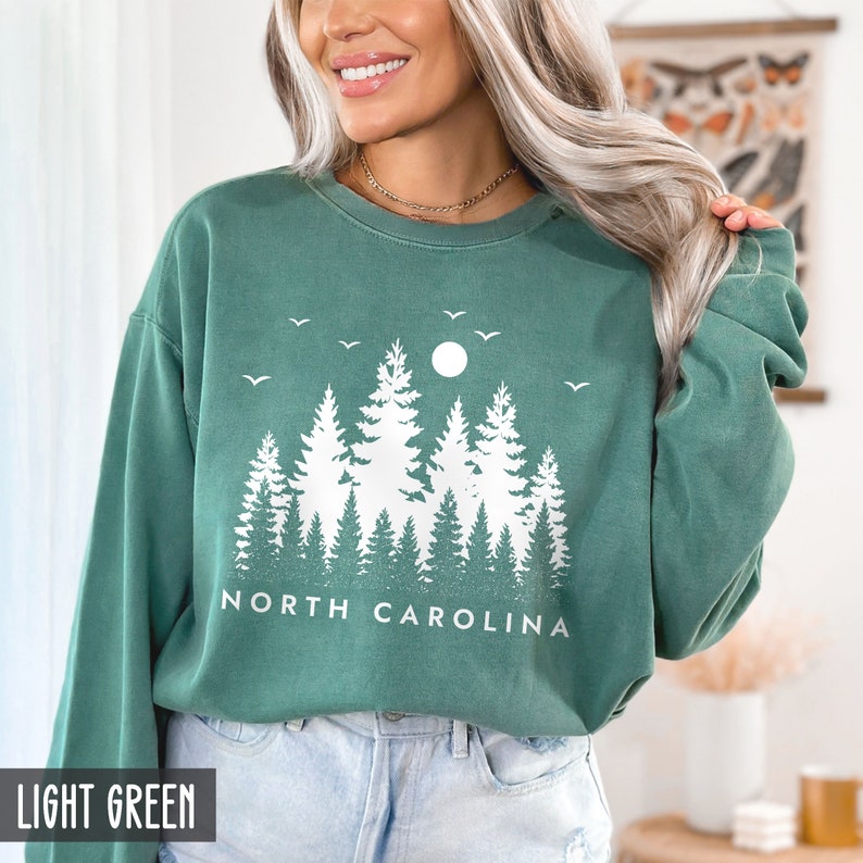 May include: A light green sweatshirt with a white graphic of a forest scene with a moon and birds. The text "NORTH CAROLINA" is printed below the graphic.