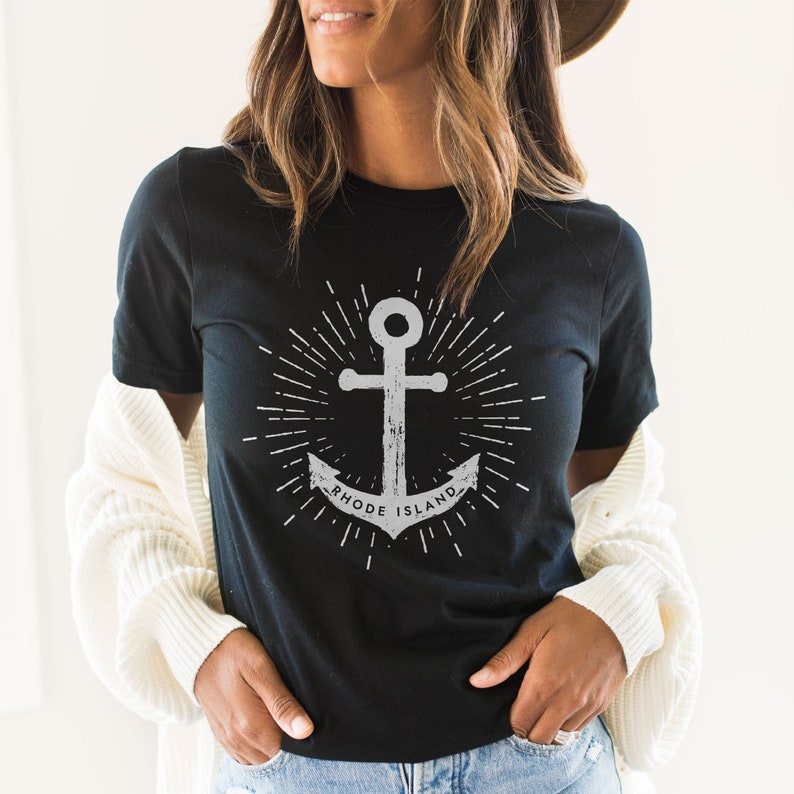 May include: Black t-shirt featuring a white anchor graphic and the text "Rhode Island" in a circle around the anchor. The anchor is surrounded by white rays of light.
