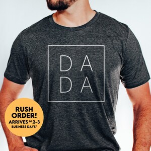 May include: A dark grey t-shirt with the word "DADA" printed in white inside a white square.