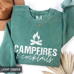 May include: Light green sweatshirt with the words "Campfires & Cocktails" in white script and a campfire graphic. A gold necklace with a pendant is on the sweatshirt. A brown hat is in the background.