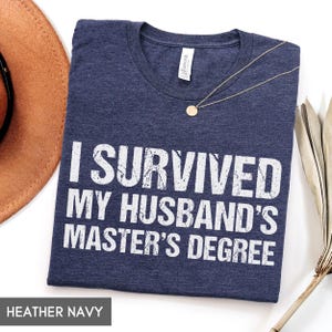 May include: Heather navy t-shirt with the text "I SURVIVED MY HUSBAND'S MASTER'S DEGREE" in white block letters. A gold necklace with a round pendant is draped on the shirt. The shirt is folded and displayed on a white surface.