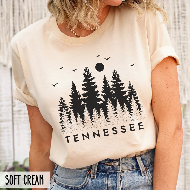 May include: A cream colored t-shirt with a black silhouette of a forest with a moon and birds. The text "TENNESSEE" is printed below the forest.