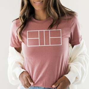 May include: A dusty rose t-shirt with a white tennis court graphic. The shirt has short sleeves and a crew neck.