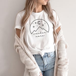 A woman wearing a white t-shirt with a black nature graphic, blue jeans, and a cream cardigan stands against a white wall.