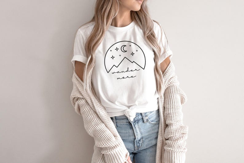 A woman wearing a white t-shirt with a black nature graphic, blue jeans, and a cream cardigan stands against a white wall.