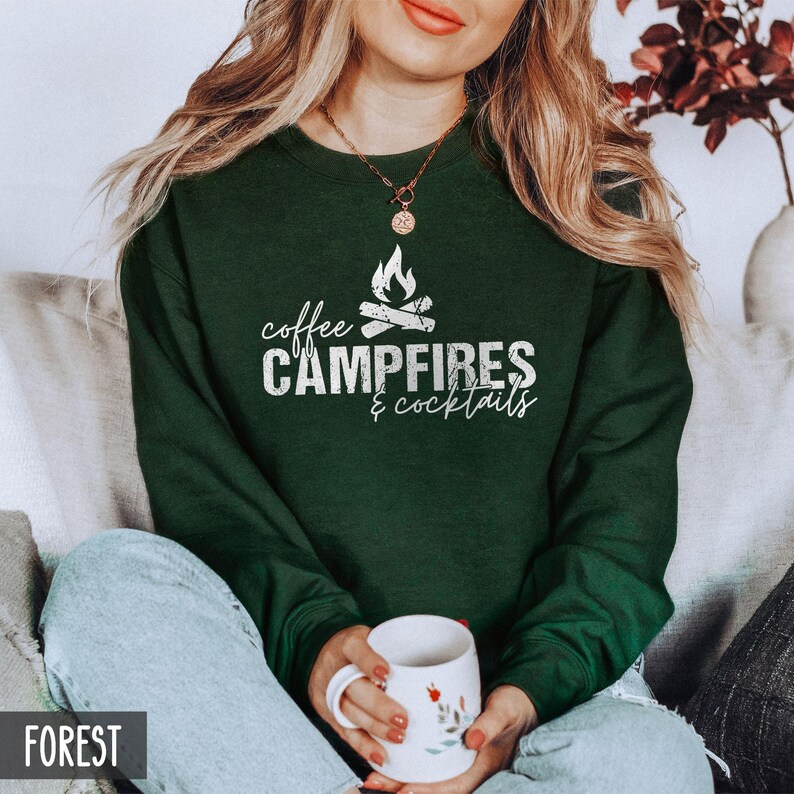 A person wears a forest green crewneck sweatshirt featuring white text that reads coffee CAMPFIRES & cocktails with a campfire graphic while holding a mug.