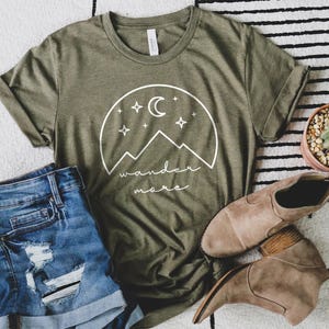 An olive green t-shirt with a white mountain graphic lies flat next to distressed denim shorts, brown ankle boots, and a small potted cactus.