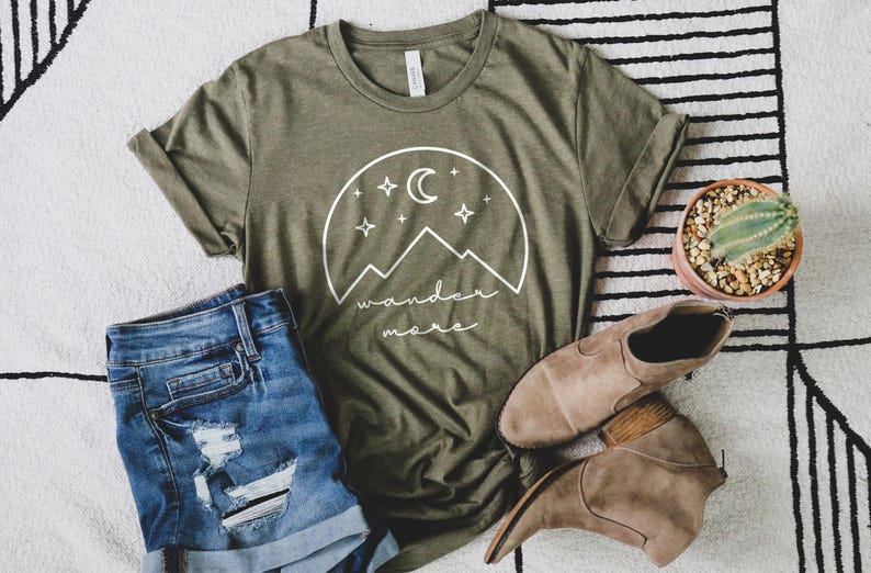 An olive green t-shirt with a white mountain graphic lies flat next to distressed denim shorts, brown ankle boots, and a small potted cactus.