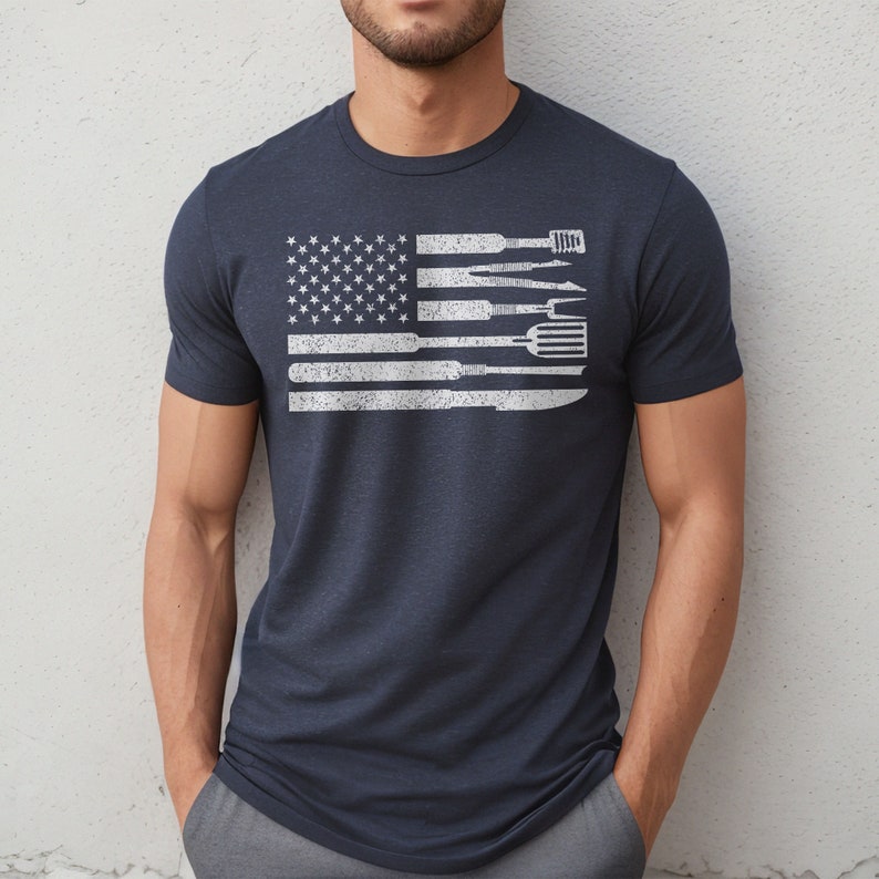 Puede incluir: Una camiseta azul marino con un dise&ntilde;o de bandera estadounidense blanca desgastada. La bandera est&aacute; compuesta por varias herramientas, incluyendo una esp&aacute;tula, un cuchillo, un tenedor y un batidor.