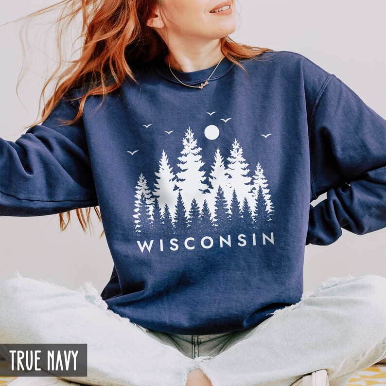 Puede incluir: Una sudadera azul marino con un gr&aacute;fico blanco de una silueta de bosque y la palabra "WISCONSIN" impresa debajo. La sudadera la lleva puesta una persona sentada con las piernas cruzadas.