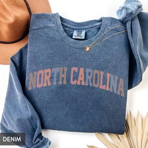 Puede incluir: Una sudadera azul vaquera con las palabras "NORTH CAROLINA" en un dise&ntilde;o de letras en bloque multicolor. Un collar dorado con un colgante est&aacute; drapeado en la parte delantera. Un sombrero marr&oacute;n es visible en el fondo.