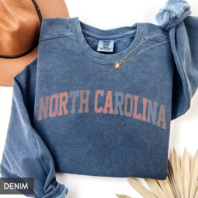 Puede incluir: Una sudadera azul vaquera con las palabras "NORTH CAROLINA" en un dise&ntilde;o de letras en bloque multicolor. Un collar dorado con un colgante est&aacute; drapeado en la parte delantera. Un sombrero marr&oacute;n es visible en el fondo.