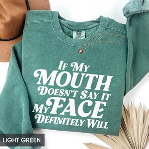 Puede incluir: Sudadera verde claro con el texto blanco "If My Mouth Doesn't Say It My Face Definitely Will". Un collar dorado con un colgante est&aacute; sobre la sudadera. Un sombrero marr&oacute;n est&aacute; en el fondo.
