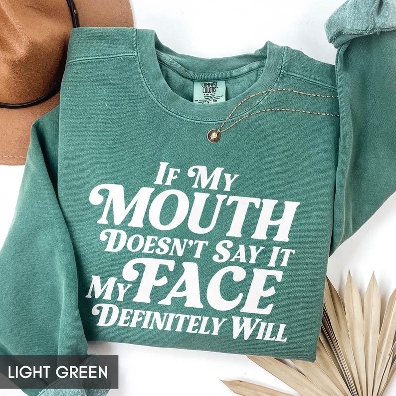 Puede incluir: Sudadera verde claro con el texto blanco "If My Mouth Doesn't Say It My Face Definitely Will". Un collar dorado con un colgante est&aacute; sobre la sudadera. Un sombrero marr&oacute;n est&aacute; en el fondo.