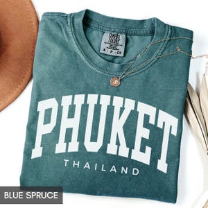 May include: Blue spruce t-shirt with the word "PHUKET" in white block letters, and "THAILAND" below. A gold necklace with a pendant rests on the shirt. The shirt is folded and has a Comfort Colors tag.