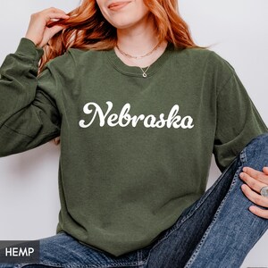 May include: An olive green sweatshirt with the word "Nebraska" in white script across the chest. The sweatshirt has long sleeves and a crew neck. The word "HEMP" is visible in the bottom left corner.