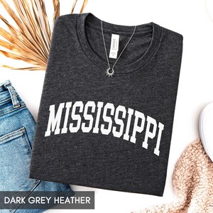 May include: A dark grey heather t-shirt featuring the word MISSISSIPPI printed in white capital letters in an arched font across the chest. The shirt has a crew neckline and a small silver sunburst pendant necklace resting on the collar area.