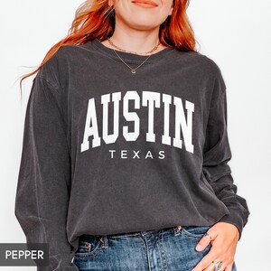 May include: A dark gray long-sleeve sweatshirt with the word "AUSTIN" in large white block letters arched across the chest, with "TEXAS" printed below. The sweatshirt is worn with blue jeans.