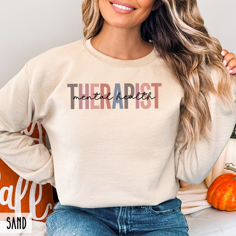 May include: A beige sweatshirt with the text "THERAPIST mental health" in colorful letters. The letters are in shades of pink, red, blue, and gray.