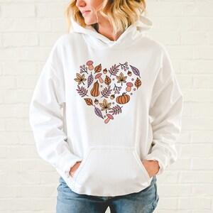 May include: White hoodie with a heart-shaped design featuring autumn-themed elements like leaves, pumpkins, acorns, and mushrooms in shades of orange, pink, and purple.