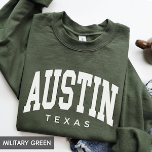 May include: Military green sweatshirt with the word "AUSTIN" in large white block letters arched across the chest, with "TEXAS" below. The sweatshirt is folded, with a portion of a denim garment visible.